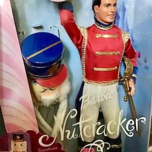 Puede incluir: Muñeca Barbie El Cascanueces con un uniforme rojo y dorado con una faja blanca y una espada. La muñeca sostiene una espada y está de pie junto a una muñeca cascanueces.