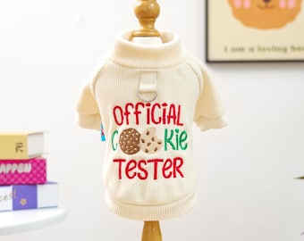 May include: Cream-colored dog sweater with a ribbed texture and a turtleneck. The front features the embroidered text "OFFICIAL COOKIE TESTER" in red and green, with cookie graphics. Includes a small loop with a metal ring.