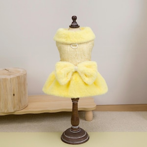 K&ouml;nnte beinhalten: Ein gelbes Hundekleid mit einem Kunstfellkragen und -rock sowie einer gro&szlig;en Schleife. Das Kleid wird auf einer Holzpuppe pr&auml;sentiert. Das Kleid hat einen D-Ring zum Anbringen einer Leine.
