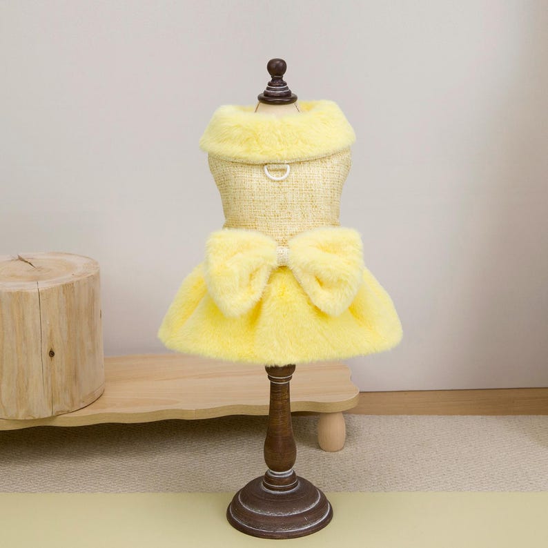 K&ouml;nnte beinhalten: Ein gelbes Hundekleid mit einem Kunstfellkragen und -rock sowie einer gro&szlig;en Schleife. Das Kleid wird auf einer Holzpuppe pr&auml;sentiert. Das Kleid hat einen D-Ring zum Anbringen einer Leine.