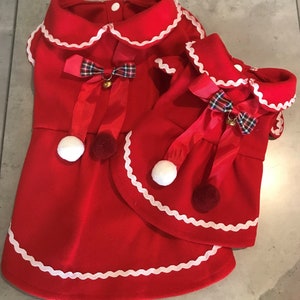May include: Two red dog dresses with white scalloped trim and red and green tartan bows. The dresses have white pom-poms and jingle bells.