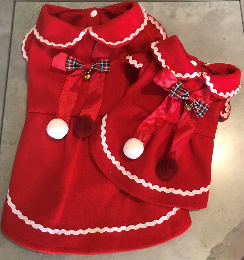 May include: Two red dog dresses with white scalloped trim and red and green tartan bows. The dresses have white pom-poms and jingle bells.