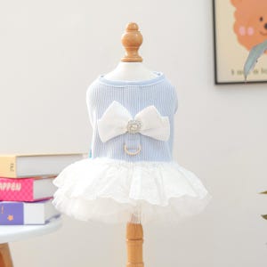 May include: A light blue dog dress with a white bow and a rhinestone center. The dress has a white lace skirt and a gold-colored metal ring for a leash. The dress is displayed on a wooden mannequin.