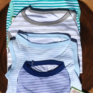 May include: Four striped tank tops for dogs in blue, white and grey colours. The tank tops are stacked on top of each other. The top tank top is turquoise and white striped. The second tank top is grey and white striped. The third tank top is light blue and white striped. The bottom tank top is dark blue and white striped.