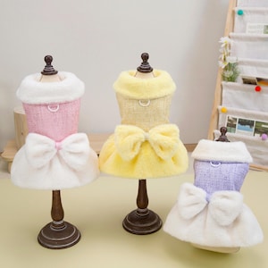 K&ouml;nnte beinhalten: Drei Hundekleider in Rosa, Gelb und Lavendel, jeweils auf einer Miniatur-Schaufensterpuppe ausgestellt. Die Kleider haben einen flauschigen Kragen, eine Schleife und einen Rock. Die Schaufensterpuppen stehen auf braunen St&auml;ndern.