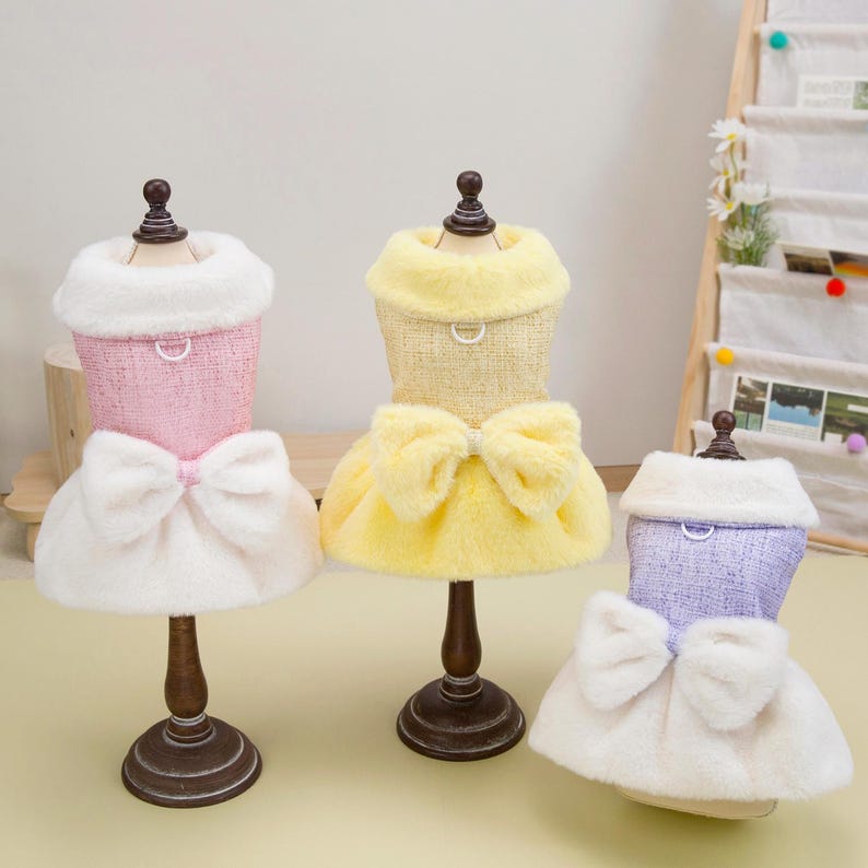 K&ouml;nnte beinhalten: Drei Hundekleider in Rosa, Gelb und Lavendel, jeweils auf einer Miniatur-Schaufensterpuppe ausgestellt. Die Kleider haben einen flauschigen Kragen, eine Schleife und einen Rock. Die Schaufensterpuppen stehen auf braunen St&auml;ndern.