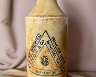 May include: A vintage, tan-colored stoneware bottle with a rounded top and a narrow neck. The bottle features a black and white triangular label with the text "HOME BREWED E ALDRIDGE & Co. GINGER BEER MINERAL WATERS SOUTHAMPTON."