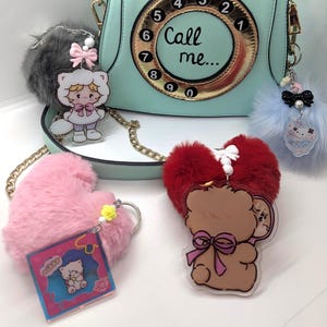 May include: A mint green crossbody bag shaped like a vintage rotary telephone with a pink handset and a gold dial featuring the text Call me. The bag is decorated with four plush pom-pom keychains in gray, light blue, pink, and red.