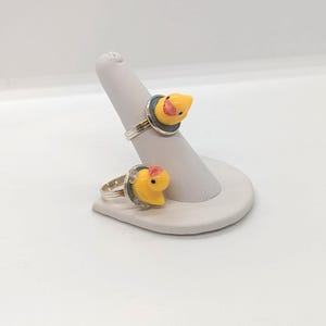 May include: Two silver-toned rings featuring small, yellow rubber duck charms with orange beaks. Each duck is mounted on a circular base. The rings are displayed on a tapered, light gray jewelry stand designed to hold rings securely in place.