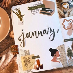 May include: A white planner page with the word "January" written in black cursive script. The page is decorated with stickers, including a record player, candles, and a woman with a bun. The page also includes the text "Holly Jolly" and "made with love".