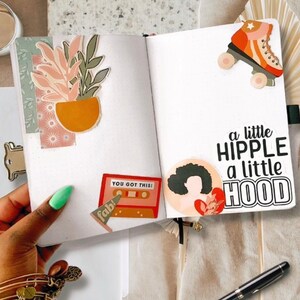 May include: A white journal with a variety of colorful stickers, including a plant, a cassette tape, a roller skate, and a woman with an afro. The text "You Got This!" and "a little hipple a little hood" are also included on the stickers.