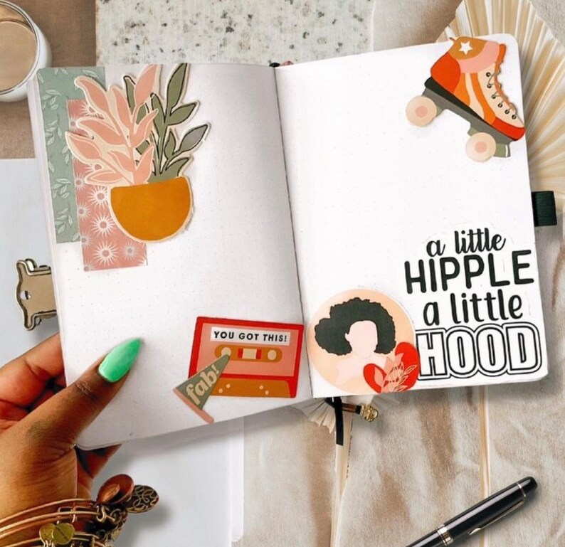 May include: A white journal with a variety of colorful stickers, including a plant, a cassette tape, a roller skate, and a woman with an afro. The text "You Got This!" and "a little hipple a little hood" are also included on the stickers.