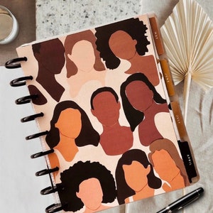 May include: A spiral bound planner with a colorful abstract design featuring multiple brown and beige human silhouettes with black hair. The planner has tabs labeled "April", "July", "June", and "May".