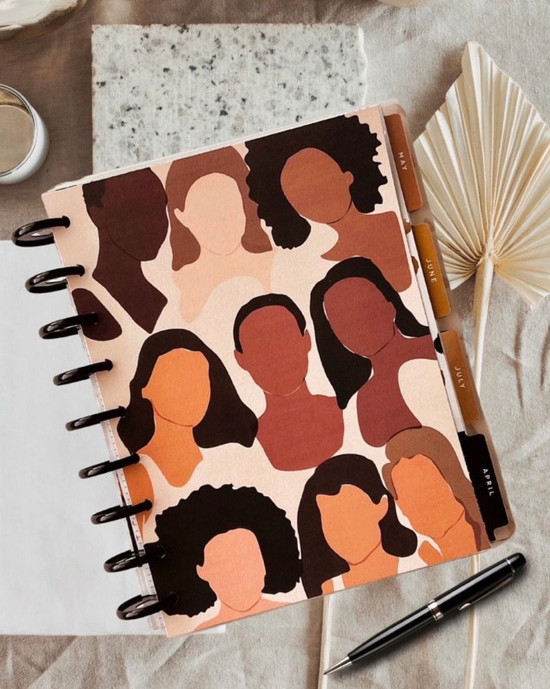 May include: A spiral bound planner with a colorful abstract design featuring multiple brown and beige human silhouettes with black hair. The planner has tabs labeled "April", "July", "June", and "May".