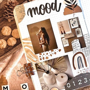 May include: A white page with the word "mood" written in black cursive. The page is decorated with brown, white, and black stickers, including a palm leaf, a rainbow, and a grid pattern. There are also several photos of people and objects, including a woman holding a branch, a vase with flowers, and a candle.