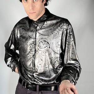 May include: A man wearing a black and silver paisley patterned shirt with a black belt and dark purple trousers.