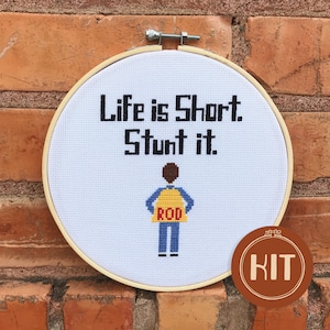 May include: A white embroidery hoop with a cross-stitch design that reads "Life is Short. Stunt it."  A cartoon figure of a person wearing a yellow vest with the word "ROD" on it is below the text. The hoop is on a brick wall with a brown circle with the word "KIT" in white text.