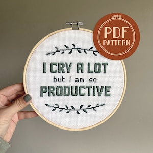 May include: A cross-stitch embroidery hoop with the text "I CRY A LOT but I am so PRODUCTIVE" in dark green. The design includes leafy accents and a brown "PDF PATTERN" sticker. The hoop is held by a hand.