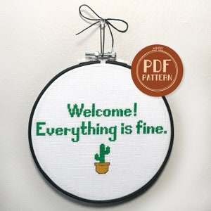 May include: A white embroidery hoop with black trim, displaying a cross-stitch design that reads "Welcome! Everything is fine." with a green cactus in a yellow pot.