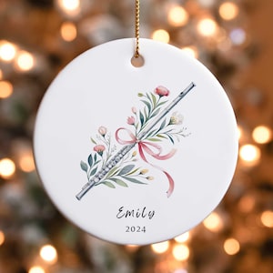 May include: A white ceramic ornament with a silver flute decorated with pink and green watercolor flowers and a pink ribbon. The ornament is personalized with the name "Emily" and the year "2024".