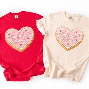 Puede incluir: Dos camisetas, una roja y otra color crema, cada una con un diseño de galleta en forma de corazón. La galleta tiene glaseado rosa, las palabras "Galentine Squad" y detalles decorativos. Las camisetas son ropa casual.