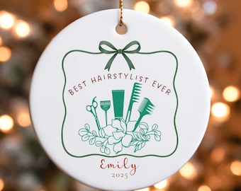 May include: White ceramic Christmas ornament with a gold string. The ornament features the text "BEST HAIRSTYLIST EVER" in dark red, along with green illustrations of hairstyling tools. The name "Emily" and the year "2025" are also included.