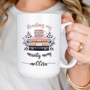 May include: White ceramic mug with a pink and blue floral design. The mug has the text "Reading my anxiety away Melissa" and a small illustration of a teacup and stack of books.