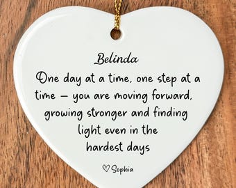 May include: White heart-shaped ceramic ornament with gold string for hanging. The ornament has the name "Belinda" and an inspirational message in black script: "One day at a time..." Signed "Sophia".