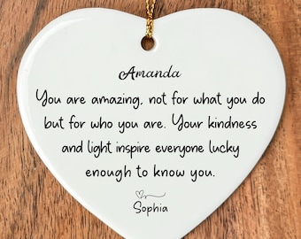 May include: White heart-shaped ceramic ornament with gold string. The ornament has the names "Amanda" and "Sophia" and a message written in black script: "You are amazing, not for what you do but for who you are. Your kindness and light inspire everyone lucky enough to know you."