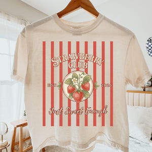 May include: Cream-colored t-shirt with a red and white striped background design. The shirt features a circular graphic with strawberries and the text "Strawberry Club" and "Just Sweet Enough."