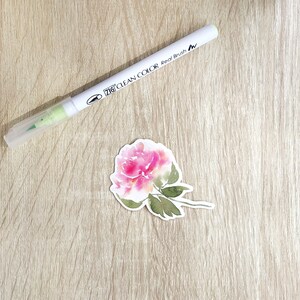 May include: A pink watercolor flower with green leaves and stems on a white background. The flower is a single bloom with a delicate, layered design. The background is a light wood grain.