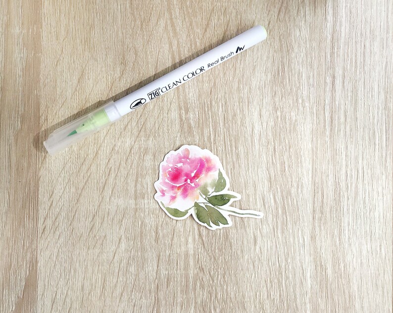 May include: A pink watercolor flower with green leaves and stems on a white background. The flower is a single bloom with a delicate, layered design. The background is a light wood grain.