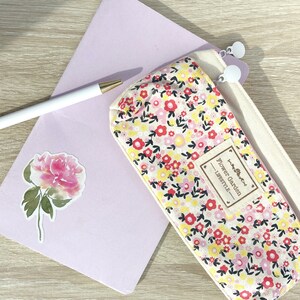 May include: A white canvas pencil case with a floral print featuring pink, red, and yellow flowers. The case has a zipper closure and a brown label that reads "Flower Garden Lifestyle".
