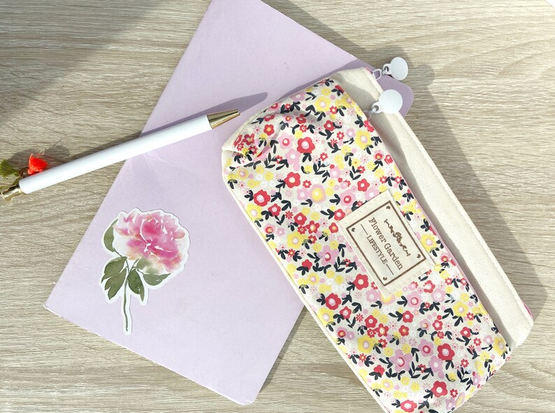 May include: A white canvas pencil case with a floral print featuring pink, red, and yellow flowers. The case has a zipper closure and a brown label that reads "Flower Garden Lifestyle".