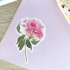 May include: A watercolor sticker of a pink and white peony flower with green leaves on a light purple background.
