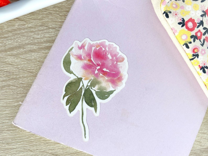 May include: A watercolor sticker of a pink and white peony flower with green leaves on a light purple background.