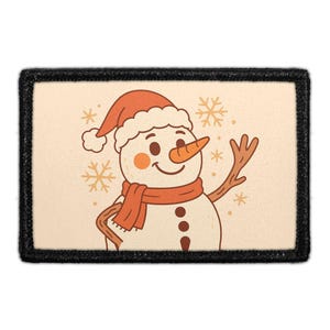 May include: A rectangular patch with a black border featuring a cartoon snowman wearing an orange hat and scarf. The snowman has a carrot nose, rosy cheeks, and three brown buttons. The background is cream-colored with orange snowflakes.
