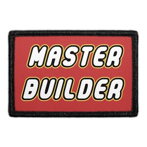 May include: A rectangular patch with a red background and a thick black border. The text MASTER BUILDER is printed in white, bold, rounded capital letters with a thin yellow outline, centered on the red field of the rectangular fabric patch.