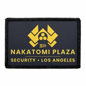 May include: A rectangular black patch with a gold embroidered border. The center features a gold geometric logo above the text NAKATOMI PLAZA, with the Japanese characters 玉砕 below the logo, and SECURITY • LOS ANGELES printed at the bottom.