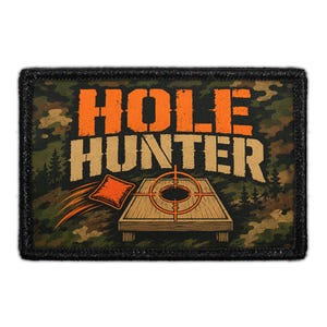 May include: A rectangular patch with a black border featuring a camouflage background. The text HOLE HUNTER is printed in orange and tan. Below the text is an illustration of a cornhole board with a target and a bean bag in mid-air.