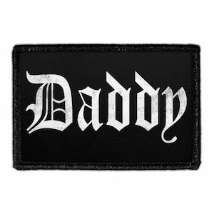 May include: A rectangular black embroidered patch featuring the word Daddy in a white, distressed, gothic-style font. The patch has a thick, stitched black border that frames the text, creating a high-contrast design with a textured appearance.