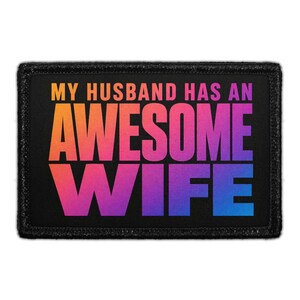 May include: A rectangular black patch with the text "MY HUSBAND HAS AN AWESOME WIFE" in a gradient of orange, pink, purple, and blue. The patch has a black border.