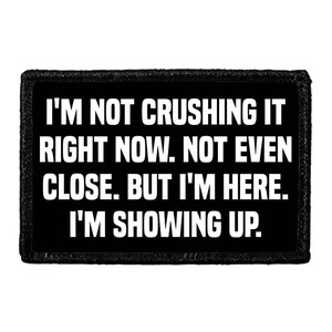 May include: A rectangular black patch with a stitched border featuring the white text: I'M NOT CRUSHING IT RIGHT NOW. NOT EVEN CLOSE. BUT I'M HERE. I'M SHOWING UP. The design is centered on a solid black background with bold, sans-serif capital letters.