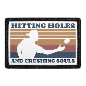 May include: A rectangular patch with a black border featuring a white silhouette of a person throwing a beanbag toward a hole. The background has horizontal stripes in tan, orange, and dark blue. The text reads HITTING HOLES AND CRUSHING SOULS.