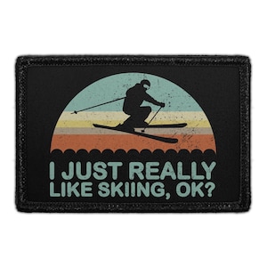 May include: A rectangular black patch featuring a distressed graphic of a skier in silhouette against a semi-circular sunset background with horizontal stripes in muted colors. The text I JUST REALLY LIKE SKIING, OK? is printed in light blue.