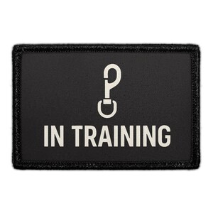 May include: A rectangular black patch with a stitched border. The center features a white graphic of a swivel snap hook icon positioned above the white text IN TRAINING in a bold, sans-serif font. The design is centered on the black fabric.