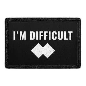 May include: A rectangular black patch with a stitched border featuring the white text I'M DIFFICULT in a bold sans-serif font, positioned above two white diamond shapes that are joined together in a horizontal orientation.