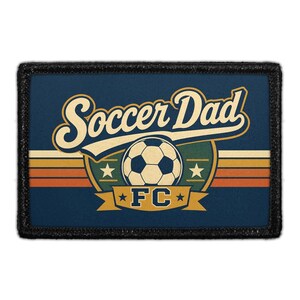 May include: A rectangular patch with a dark blue background featuring the text Soccer Dad in cream script. Below, a soccer ball sits above a gold banner with the text FC. The design includes horizontal stripes in orange, yellow, and cream.