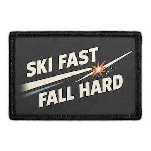 May include: A rectangular black patch with a stitched border featuring the white text "SKI FAST" and "FALL HARD" separated by a graphic of a white streak ending in an orange and white starburst explosion on a dark gray textured background.