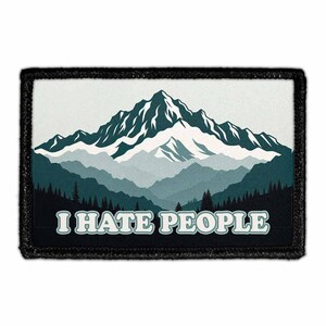 May include: A rectangular embroidered patch with a black border featuring a mountain range illustration in shades of teal, blue, and white. Below the mountains, a dark forest silhouette sits above the white text I HATE PEOPLE.